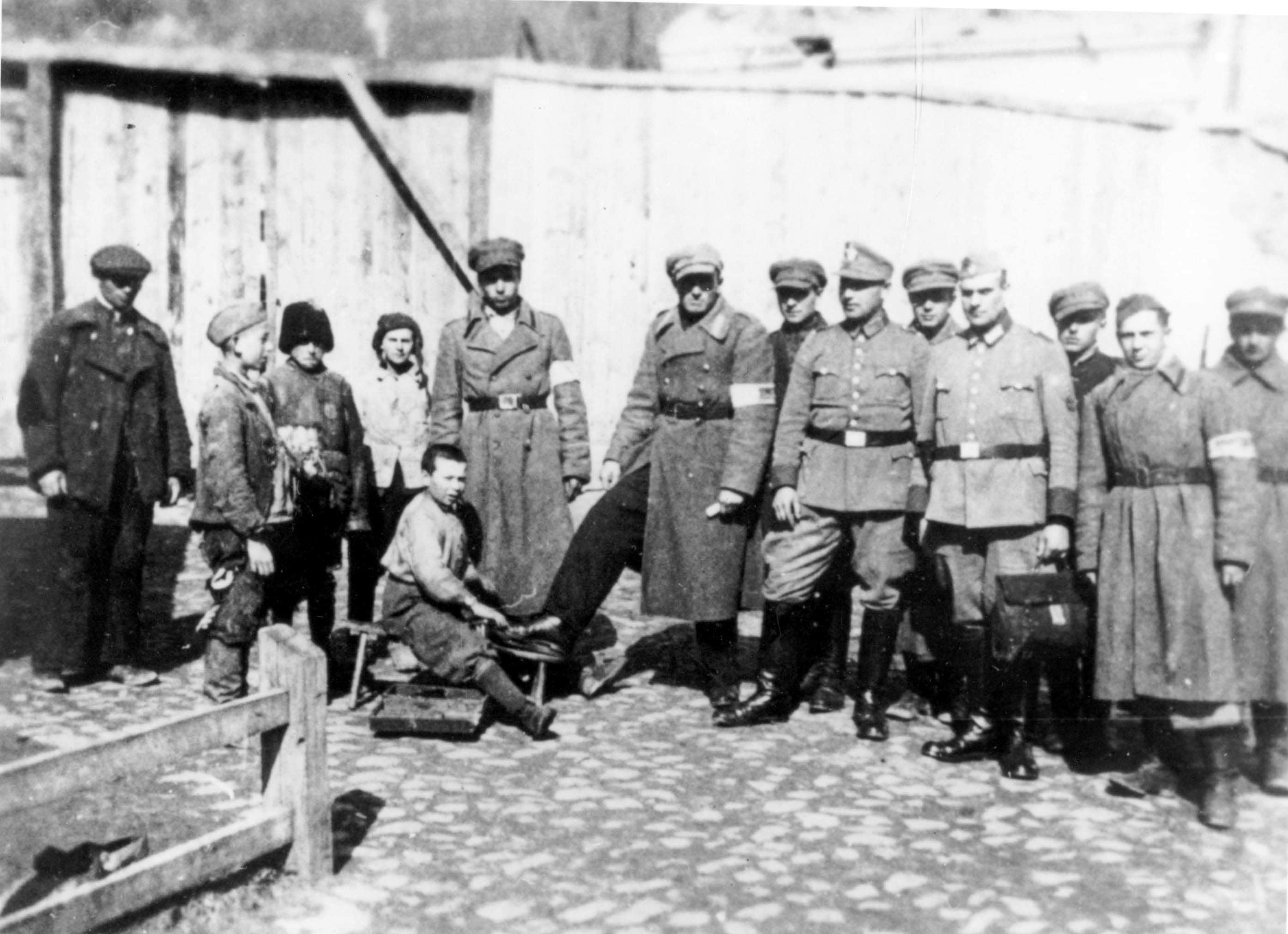 At the main gate of the Krzemieniec ghetto, Jewish boy polishing the boots of Jewish policemen as German policemen look on